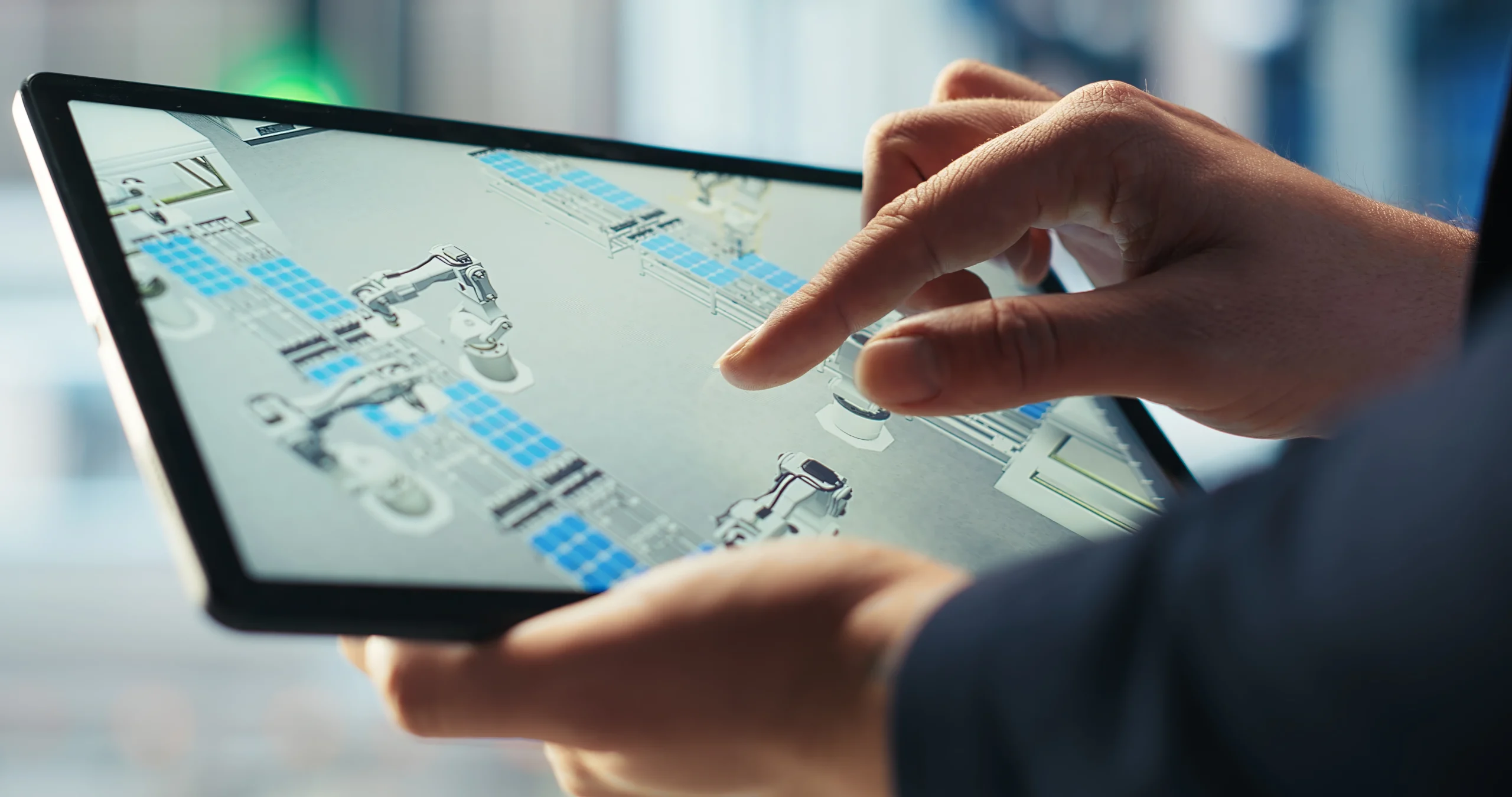 Close-up of a person's hands using a tablet to monitor an automated factory floor. The screen displays a top-down digital twin of a production line where robotic arms are assembling blue solar panels.