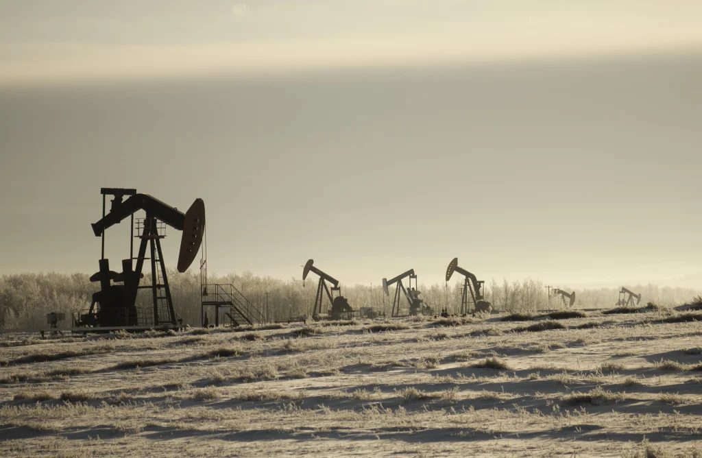 Silhouette of two oil pumpjacks (nodding donkeys) operating in a field against a bright, hazy sky.