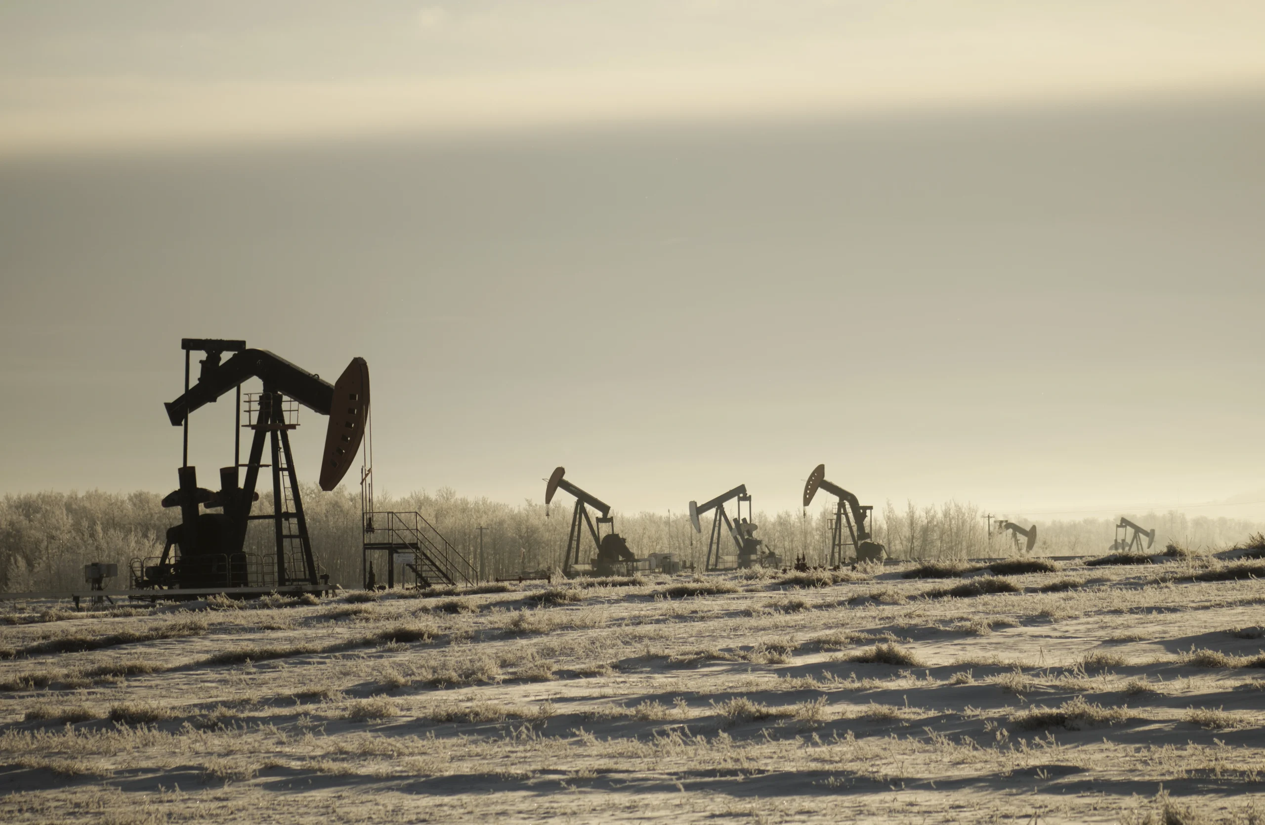 Silhouette of two oil pumpjacks (nodding donkeys) operating in a field against a bright, hazy sky.